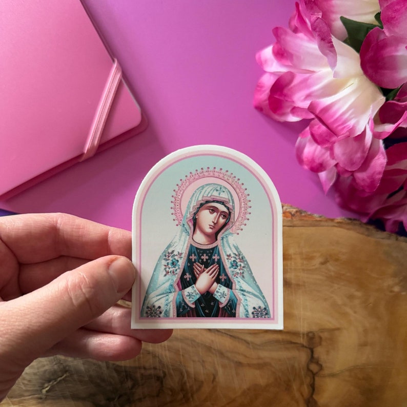 May include: A sticker featuring a religious icon of the Virgin Mary. The image is in a blue, pink, and white color scheme. The Virgin Mary is depicted with her hands clasped in prayer, wearing a blue robe and a white veil.