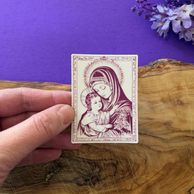 May include: A sticker with a vintage illustration of the Virgin Mary holding baby Jesus. The image is in shades of pink and white on a cream background.