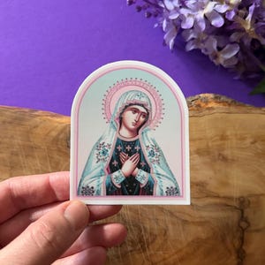 May include: A sticker featuring a religious icon of the Virgin Mary. The image is in a traditional style with a blue and white robe and a pink halo. The sticker is on a white background.