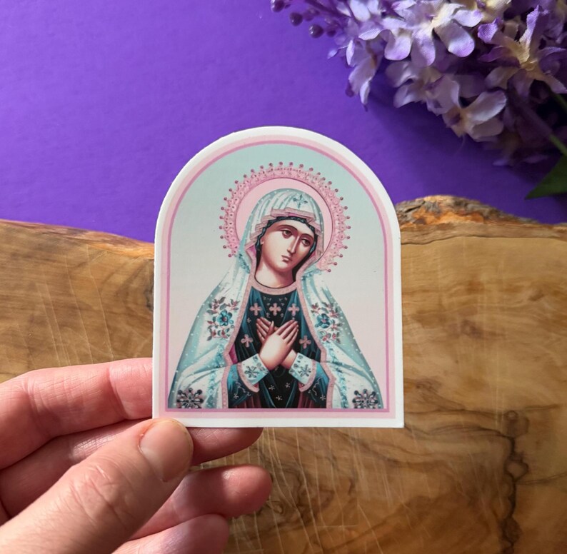 May include: A sticker featuring a religious icon of the Virgin Mary. The image is in a traditional style with a blue and white robe and a pink halo. The sticker is on a white background.
