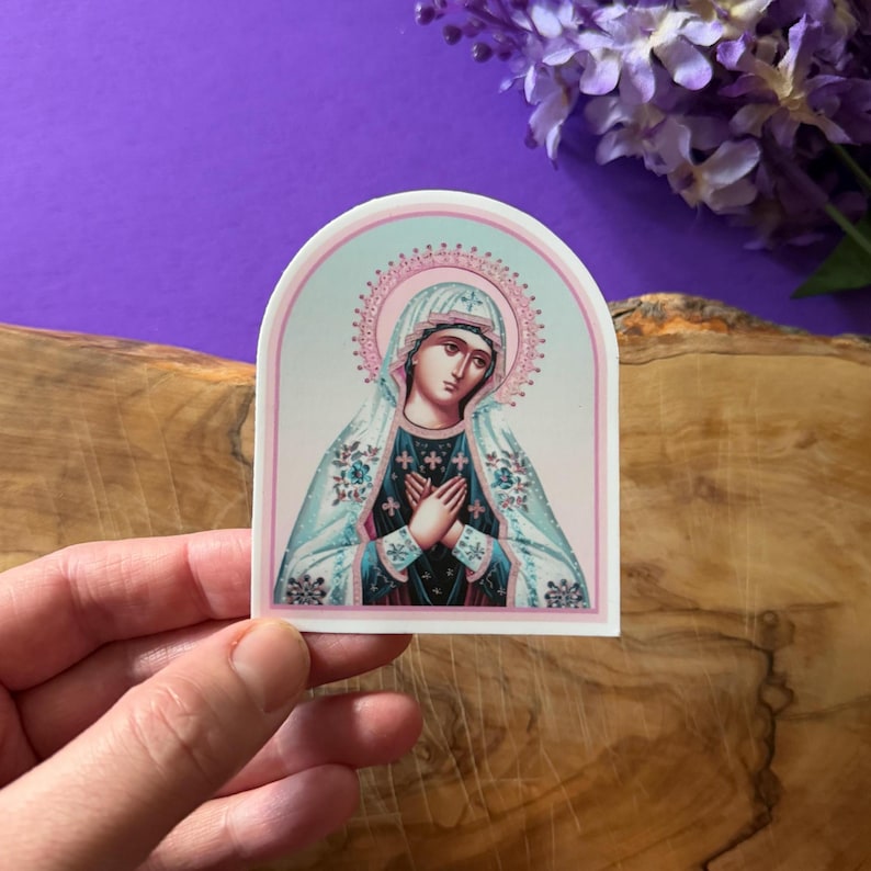May include: A sticker featuring a religious icon of the Virgin Mary. The image is in a vintage style with a blue and pink color scheme. The Virgin Mary is depicted with her hands clasped in prayer and wearing a blue and white robe.