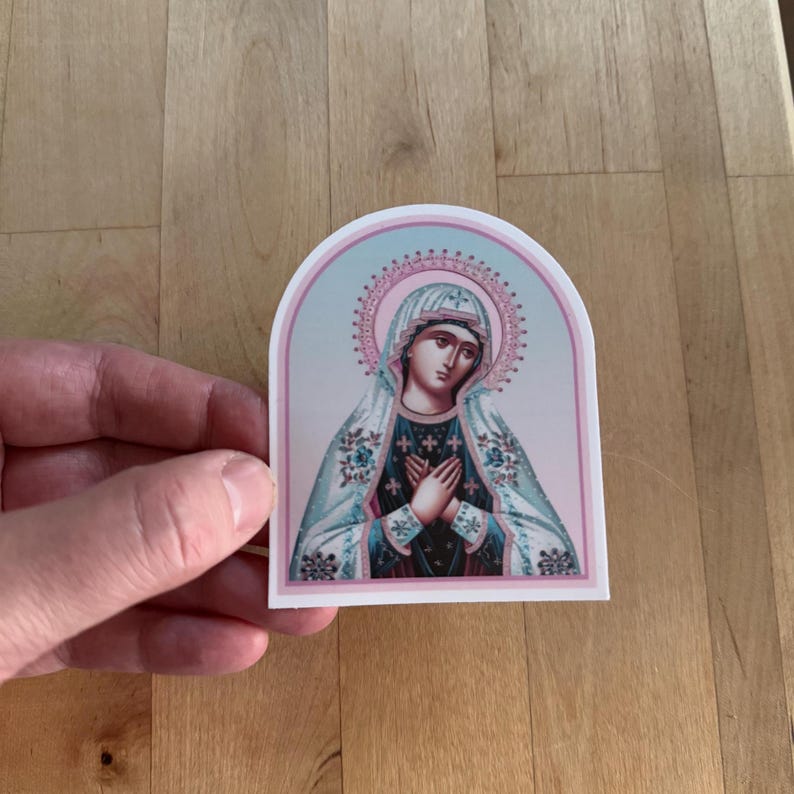 May include: A sticker featuring a religious icon of the Virgin Mary. The image is in a vintage style with a blue and pink color scheme. The Virgin Mary is depicted with her hands clasped in prayer and wearing a white veil.