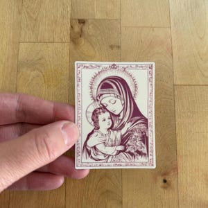 May include: A rectangular sticker featuring a religious illustration of a woman in a head covering holding a child. The image is in shades of burgundy and cream, with intricate border details and a halo around the woman's head.