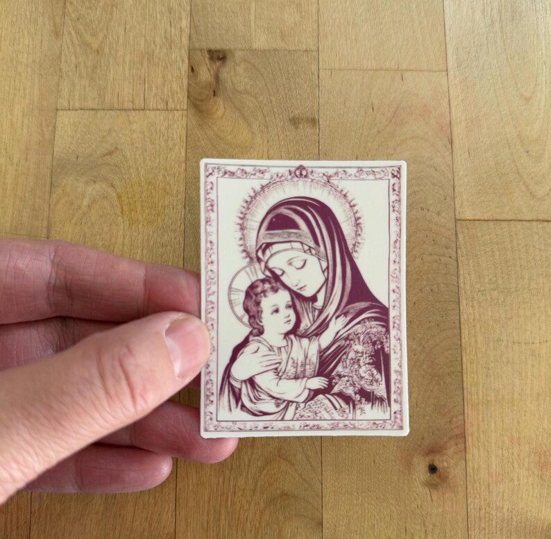 May include: A rectangular sticker featuring a religious illustration of a woman in a head covering holding a child. The image is in shades of burgundy and cream, with intricate border details and a halo around the woman's head.