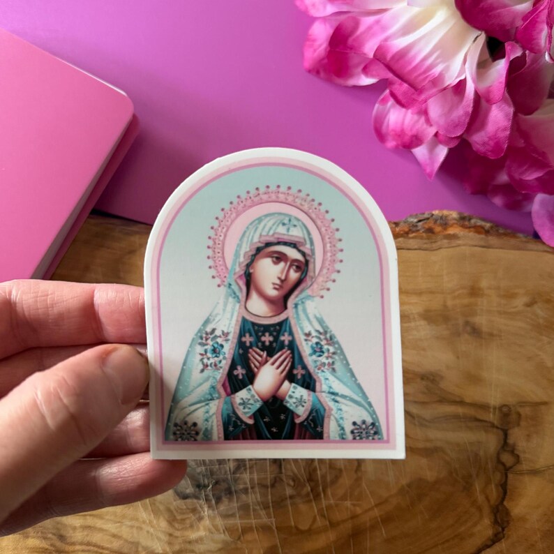 May include: A sticker featuring a religious icon of the Virgin Mary in a blue and white robe with a pink halo. The sticker is on a white background with a light blue and pink border.