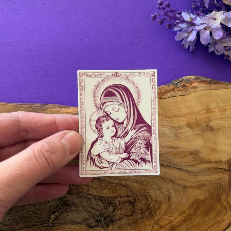 May include: A rectangular sticker featuring a religious image of a woman and child in a purple and cream color scheme. The woman wears a head covering and holds the child. The image has a halo and is set against a cream background.