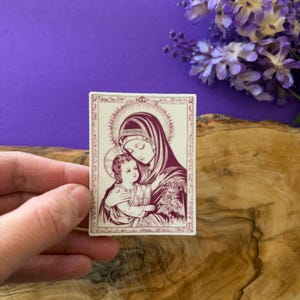 May include: A rectangular sticker featuring a religious illustration of Mary and Jesus in a burgundy and cream color scheme. The image depicts Mary holding Jesus, with a halo around their heads. The sticker has a decorative border.