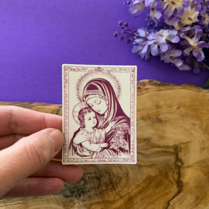 May include: A rectangular sticker featuring a religious illustration of Mary and Jesus in a burgundy and cream color scheme. The image depicts Mary holding Jesus, with a halo around their heads. The sticker has a decorative border.