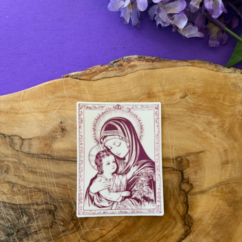 May include: A rectangular sticker featuring a religious illustration of a mother and child in shades of burgundy and white. The image is set against a wooden surface with purple flowers in the background.