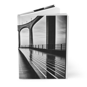 May include: A black and white photograph of a bridge with a long, narrow walkway. The walkway is made of concrete and has a metal railing on either side. The bridge is made of concrete and has a series of arches. The photograph is taken from a low angle, looking up at the bridge.