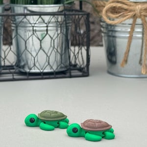 May include: Two small plastic turtle figurines. One is green with a brown shell, and the other is brown with a brown shell. They are sitting on a light gray surface.