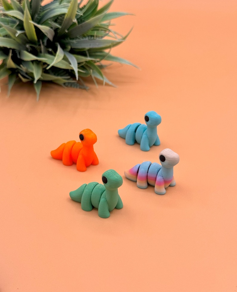 May include: Four small, segmented dinosaur figurines in different colors. The dinosaurs are orange, green, blue, and a rainbow gradient. The dinosaurs are posed in a standing position with their heads tilted up.