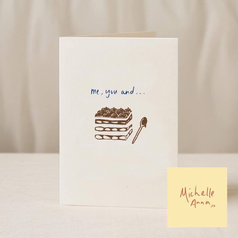 May include: A cream-coloured greeting card with a brown illustration of a layered dessert, a spoon, and the text "me, you and...". A small yellow sticky note with the name "Michelle Anna" is in the lower right corner of the card.