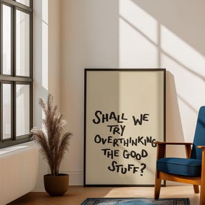 May include: A framed poster with the text "Shall we try overthinking the good stuff?" stands on a wooden floor. A blue armchair and a decorative plant in a brown pot are also visible. A blue rug is in front of the poster.