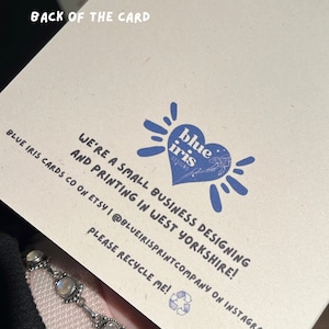 May include: Back of a card with a blue heart logo that says "blue iris designs". The text reads "We're a small business designing and printing in West Yorkshire! Blue Iris Cards co on Etsy | @blueirisprintcompany on Instagram. Please recycle me!"