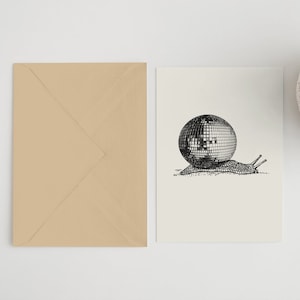 May include: A greeting card featuring a black and white illustration of a snail with a disco ball shell. A tan envelope is to the left of the card. A small succulent plant in a white pot is in the upper right corner.