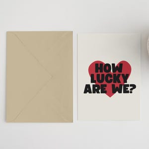 Pu&ograve; includere: Un biglietto bianco con un cuore rosso e testo nero che dice "How lucky are we?" Il biglietto &egrave; su una superficie bianca con una busta marrone e una pianta in vaso.
