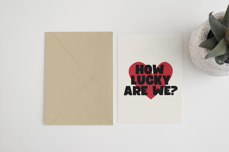 Pu&ograve; includere: Un biglietto bianco con un cuore rosso e testo nero che dice "How lucky are we?" Il biglietto &egrave; su una superficie bianca con una busta marrone e una pianta in vaso.