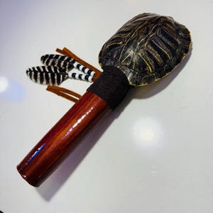 May include: A handcrafted rattle featuring a turtle shell top, a dark brown wooden handle, and a black fabric band. Decorative feathers with black and white stripes and brown leather accents are attached to the side. The rattle is designed for musical purposes.