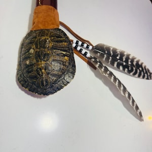 May include: A rattle featuring a dark brown wooden handle attached to a patterned turtle shell. The shell is secured with tan suede leather and decorated with two long, striped feathers attached by leather cords with black and white beads.