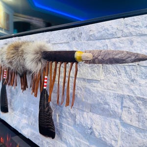 May include: A decorative spear featuring a sharp, grey stone arrowhead attached to a wooden shaft. The shaft is wrapped in dark brown leather, adorned with fluffy tan and grey fur, long brown leather fringe, and black feathers with red and white beads.