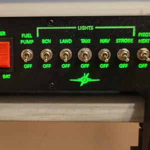以下が含まれることがあります： 緑色の文字と赤いスイッチが付いた黒いコントロールパネル。キー操作による始動、"Fuel Pump"、"Lights"、"Avionics"とラベル付けされたトグルスイッチを備えています。"Master"、"Alt"、"Bat"、"Bus 1"、"Bus 2"のテキストが含まれています。