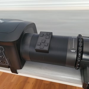 May include: A black device with multiple buttons and dials. The device has a cylindrical body with a control panel and a small rectangular button pad attached. The device is on a white surface.
