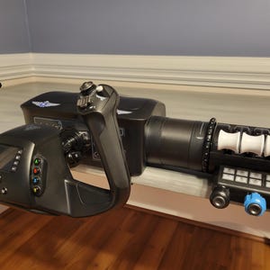 May include: A black flight simulator yoke with a small screen, buttons, and a throttle control. The yoke has a white wing design on the top. The throttle control has a series of white rollers and a panel of buttons.