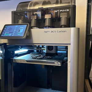 Peut inclure: Imprimante 3D Bambu Lab X1-Carbon argent et noir avec une unité AMS attachée. L'imprimante dispose d'un écran tactile affichant les données de fonctionnement et d'un plateau d'impression avec un objet imprimé. L'unité AMS contient plusieurs bobines de filament.