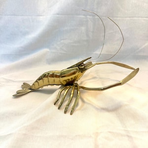 May include: A brass shrimp sculpture with detailed legs, antennae, and tail fins. The shrimp is posed on a white surface. The sculpture is made of shiny, gold-colored metal.
