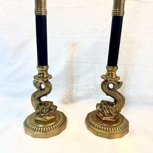 May include: A pair of antique brass candlesticks with black shafts. Each candlestick has a dolphin base and a brass top. The candlesticks are displayed on a white surface.