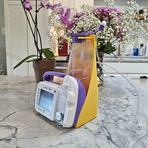 May include: A medical device with a white and purple pump, a clear fluid bag, and yellow holder. The device has a digital display and clear tubing. The background includes flowers in a vase and a potted plant.