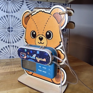 May include: A wooden G-Tube Station in the shape of a bear. The bear has a light brown face and ears, with a blue and white device in the center. The device has the words "G-Tube Station" and a screen. Clear tubing is attached.