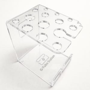 May include: Clear acrylic G-Tube Station with multiple circular openings for holding items. The stand has a rectangular top and a base with the text "G-Tube Station". The stand is designed to hold multiple items.