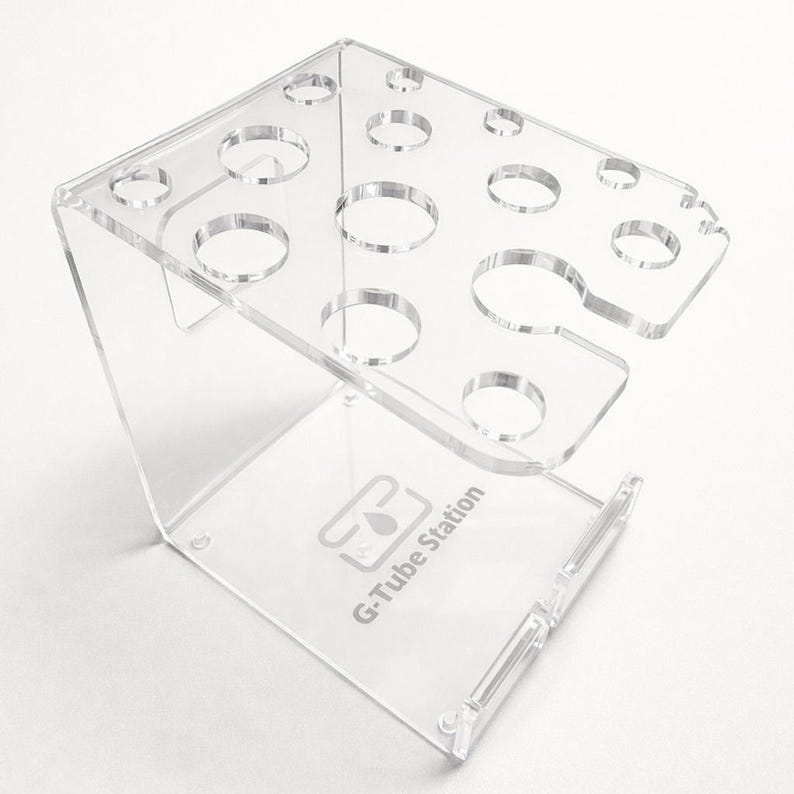May include: Clear acrylic G-Tube Station with multiple circular openings for holding items. The stand has a rectangular top and a base with the text "G-Tube Station". The stand is designed to hold multiple items.