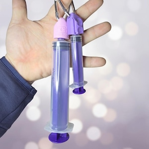 Puede incluir: Dos jeringas de plástico transparente y morado con detalles blancos y morados están unidas a un llavero plateado. Las jeringas tienen un émbolo y una base. El fondo es un bokeh borroso de blanco y morado.