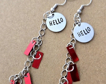 Op de afbeelding: Een paar bungelende oorbellen met zilverkleurige metalen haken en kettingen. Elke oorbel bevat een ronde zilveren bedel met het woord HELLO in zwarte hoofdletters, geaccentueerd door meerdere kleine, rechthoekige rode metalen hangertjes.