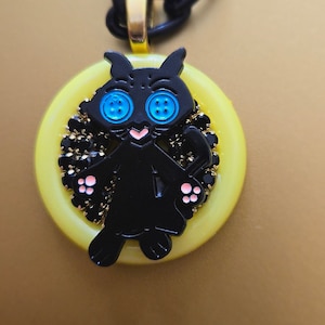 May include: A black cat charm necklace with blue eyes and pink paws. The charm is set in a yellow button-shaped pendant with black rhinestones around the edge.