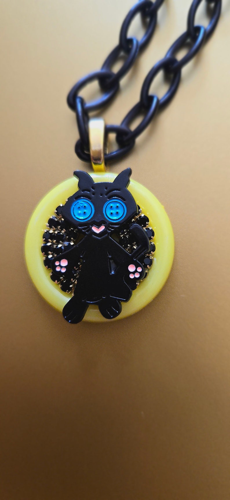May include: A black cat charm necklace with blue eyes and pink paws. The charm is set in a yellow button-shaped pendant with black rhinestones around the edge.