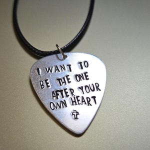 May include: A silver-colored guitar pick pendant with the words "I WANT TO BE THE ONE AFTER YOUR OWN HEART" stamped on it. A small cross is stamped at the bottom. The pendant hangs from a black cord necklace.