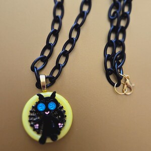 May include: A black chain necklace with a yellow button featuring a black cat with blue eyes. The cat is surrounded by black glitter.