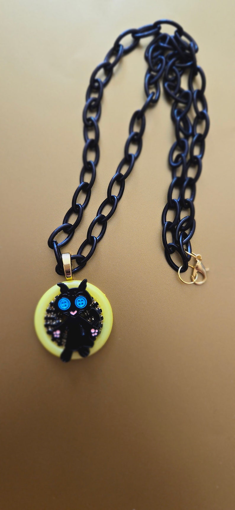 May include: A black chain necklace with a yellow button featuring a black cat with blue eyes. The cat is surrounded by black glitter.