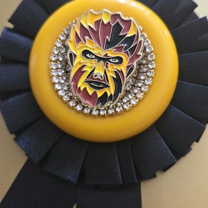 May include: A yellow and black rosette with a gold-rimmed circular pin featuring a colorful, stylized face with a beard. The pin is surrounded by a ring of clear rhinestones.