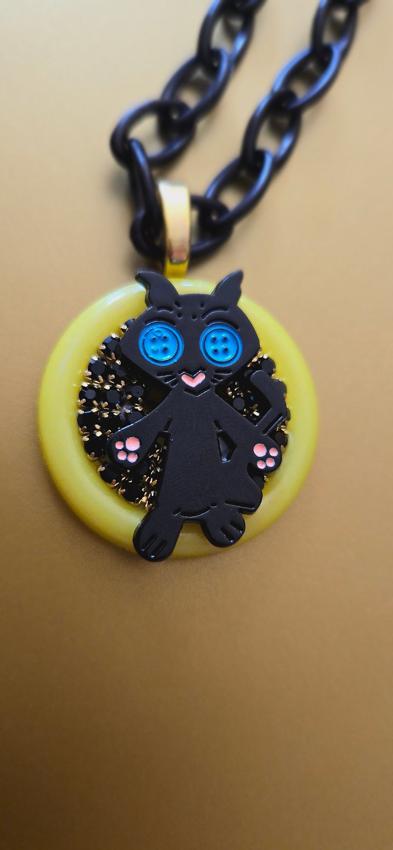 May include: A black cat charm necklace with blue button eyes and pink paws. The charm is set in a yellow circle with black beads around the edge. The necklace is made of a black chain.
