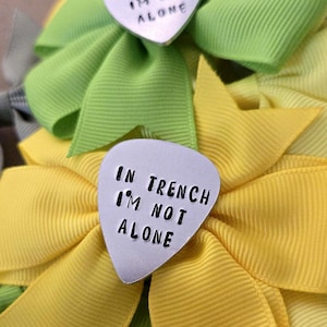 May include: A silver-toned metal guitar pick featuring the stamped black text IN TRENCH I'M NOT ALONE, centered on a bright yellow fabric bow with ribbed texture. A second identical guitar pick is visible on a lime green fabric bow behind it.