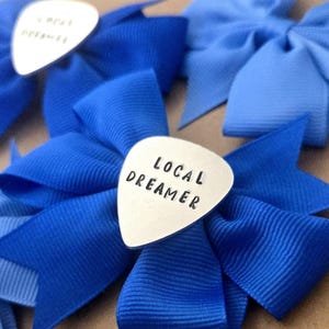 May include: A collection of blue fabric bows, each featuring a silver-toned metal guitar pick attached to the center. The metal picks are stamped with the black text LOCAL DREAMER. The bows vary in shades of blue, ranging from light to royal blue.