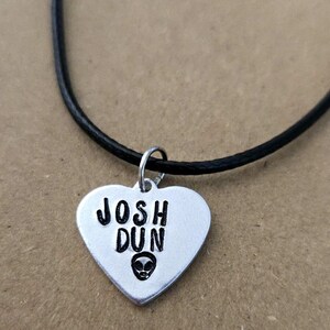 May include: A silver-toned heart-shaped pendant on a black cord necklace. The pendant is stamped with the black text JOSH DUN and features a small black alien head icon centred below the name. The metal charm hangs from a simple silver jump ring.