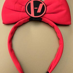 May include: A bright red headband with a large bow and a circular emblem. The emblem is black with a red design in the center. The headband is red with black accents at the ends.