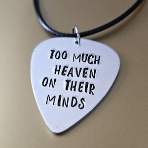 May include: A silver guitar pick pendant with the phrase "TOO MUCH HEAVEN ON THEIR MINDS" stamped in black lettering. The pendant hangs from a black cord necklace.