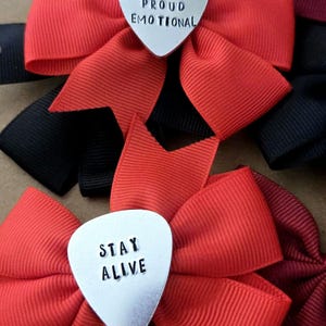 May include: A collection of fabric hair bows in red, black, and maroon, each featuring a silver-toned metal guitar pick center. One pick is stamped with the text FEW PROUD EMOTIONAL, and another pick is stamped with the text STAY ALIVE.
